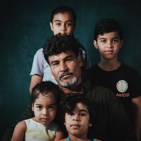 A man with a beard and dark hair, sits surrounded by four young children.