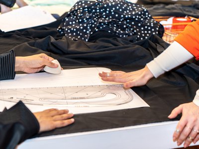 A woman drafts a pattern on top of black fabric, with another woman reviewing her work.