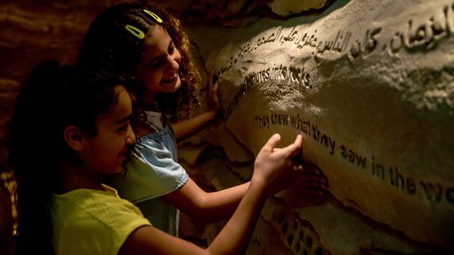 طفلان مبتسمان يكتشفان المنحوتات الصخرية على الجدار في ملعب كهف العجائب في متحف قطر الوطني