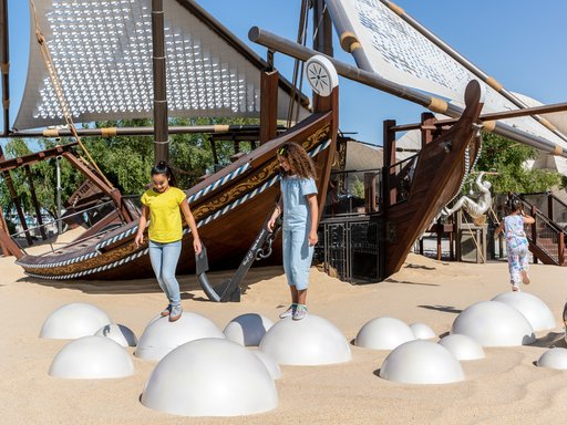 Two children in NMoQ's playground standing on top of round silver balls sunken into sand with two large wooden boats (dhows) in the background