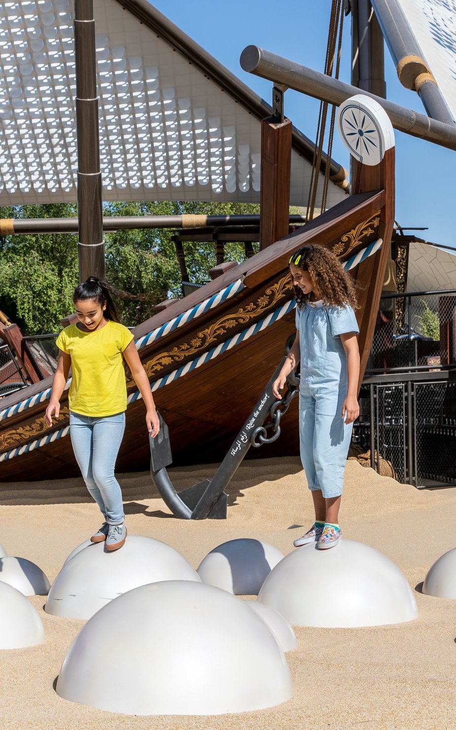 Two children in NMoQ's playground standing on top of round silver balls sunken into sand with two large wooden boats (dhows) in the background