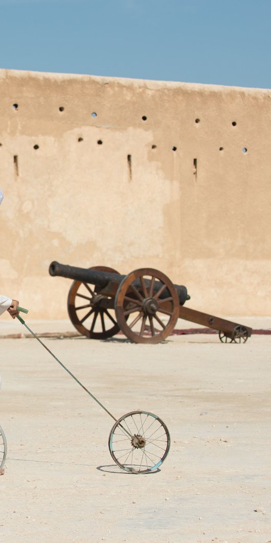 Two boys dressed in traditional Qatari attire hoop rolling with Al Zubarah fort in the background