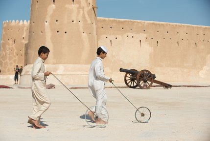 Two boys dressed in traditional Qatari attire hoop rolling with Al Zubarah fort in the background