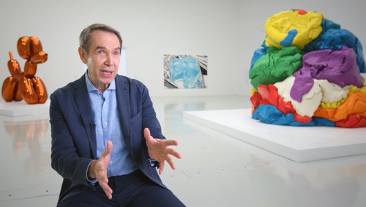 Jeff Koons at his exhibition, behind him are his public art displays of a orange balloon dog on the left and a play dough on the right