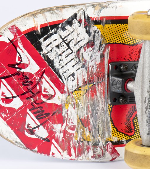 A close up of the underbelly of a skateboard, featuring red, black and yellow stickers and a black felt-tipped Tony Hawk signature.