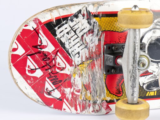 A close up of the underbelly of a skateboard, featuring red, black and yellow stickers and a black felt-tipped Tony Hawk signature.