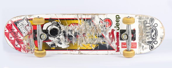 Underbelly of a skateboard is shown with scratched stickers, yellow wheels, and a felt-tipped Tony Hawk signature at the nose.