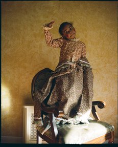 A young girl stands on an antique chair, while she plays 'dress-up'.