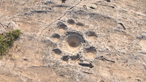A sand coloured rock face with a single large circular bowl shape carved into the rock with nine smaller circular shapes running around the outside