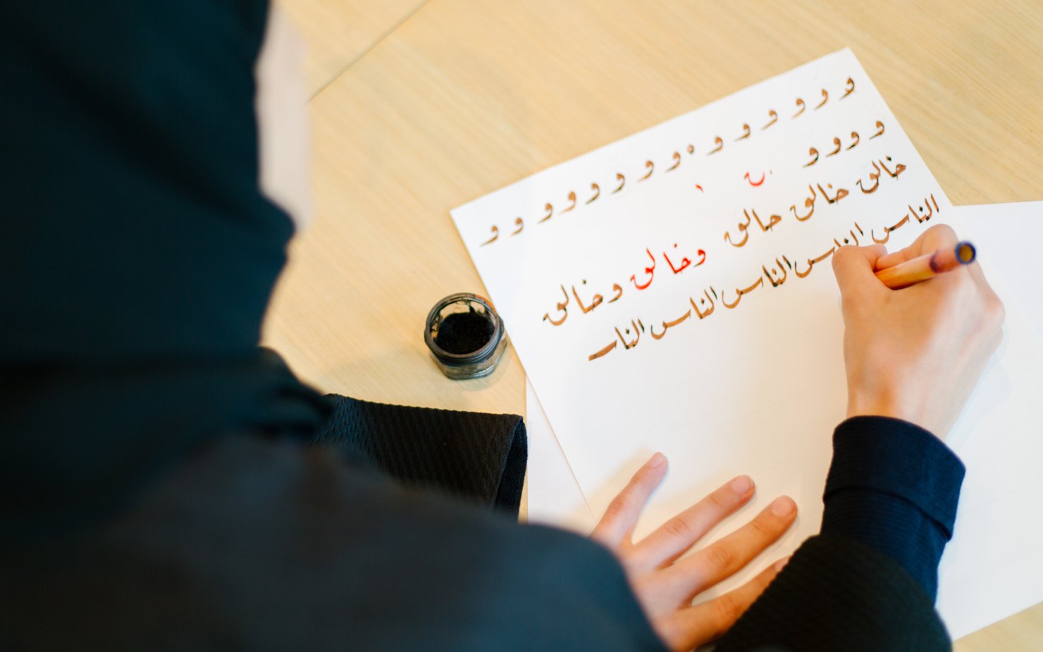 An over-the-shoulder shot of a woman in traditional Qatari clothes writing calligraphic letters on a piece of paper