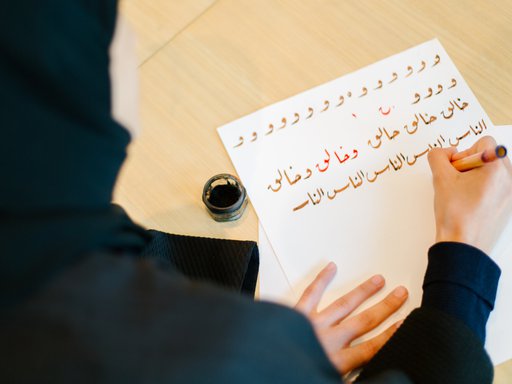 An over-the-shoulder shot of a woman in traditional Qatari clothes writing calligraphic letters on a piece of paper
