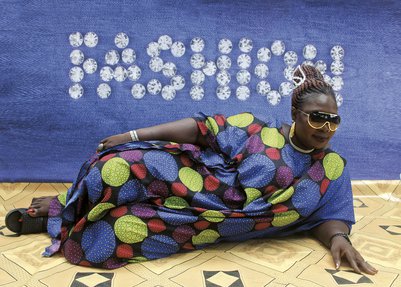 A woman lays her side on the floor, wearing a loose pattered dress and sunglasses, against a backdrop with the word 'fashion'.