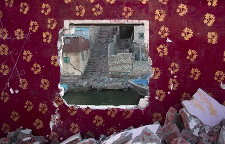 View of an urban river bank with buildings, boat and stairway, seen through a hole in a wall of a derelict building.