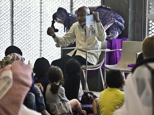 أطفال يجلسون للاستماع إلى قصة بينما يحمل أمين المكتبة كتابًا صغيرًا ويشير إلى الأطفال