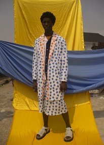 A person wears patterned clothing, standing in front of a background of outstretched yellow and blue fabrics.