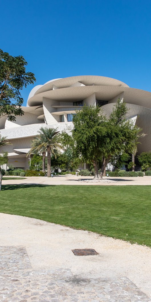 لقطة لحديقة المتحف الإسلامي مع الأشجار والعشب والممرات ويظهر متحف قطر الوطني في الخلفية.