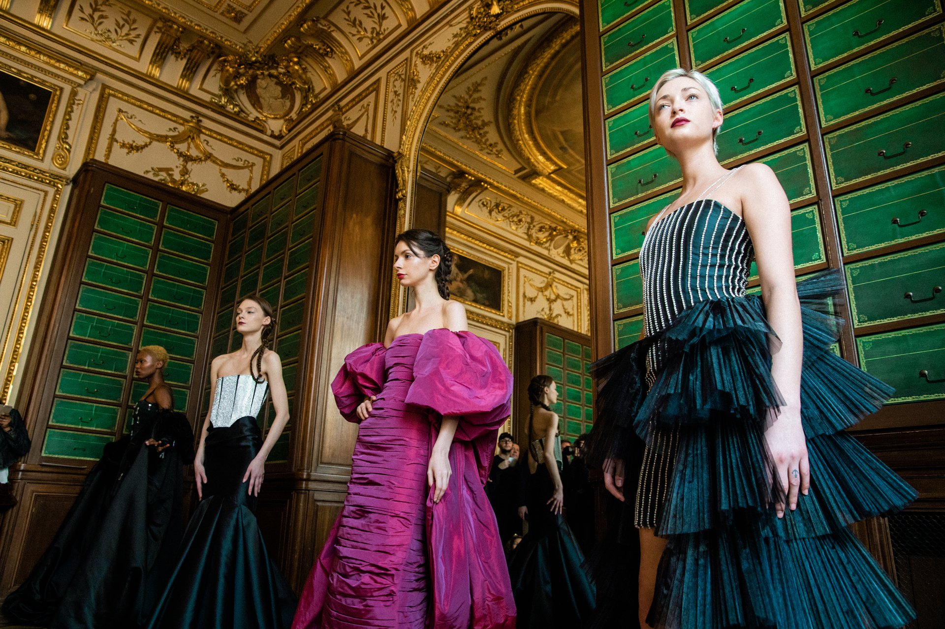 Four female fashion models wearing evening dresses in an ornate room.