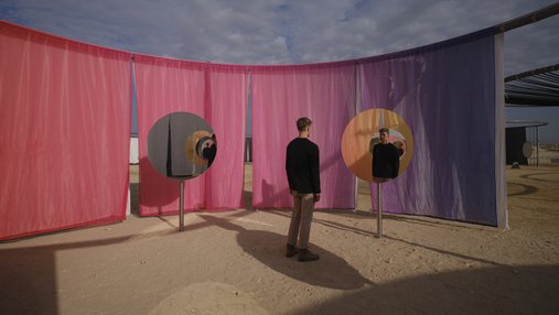 A man standing in a desert landscape looking into a round mirror with pink and purple curtains in the background