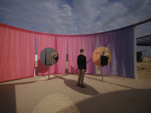 A man standing in a desert landscape looking into a round mirror with pink and purple curtains in the background